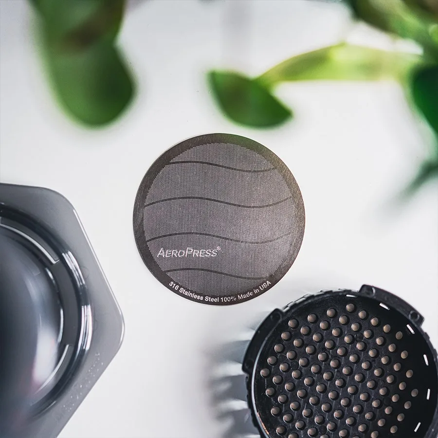 Filtro metálico estándar AeroPress junto a una cafetera y su tapa