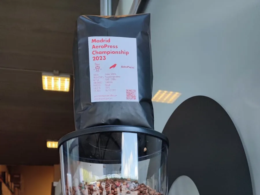 Molino con granos en la tolva y una bolsa negra «Madrid AeroPress Championship 2023» encima.