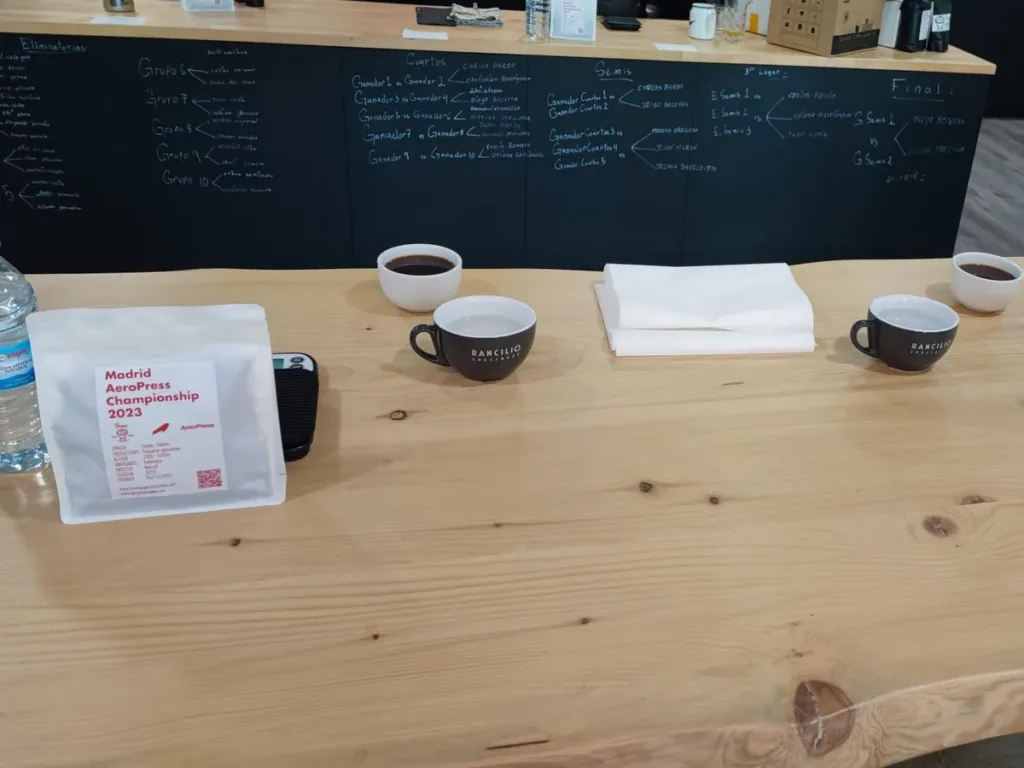 Mesa de madera con tazas de cata y dos bolsas blancas del campeonato, delante de una pizarra con el cuadro de la competición.