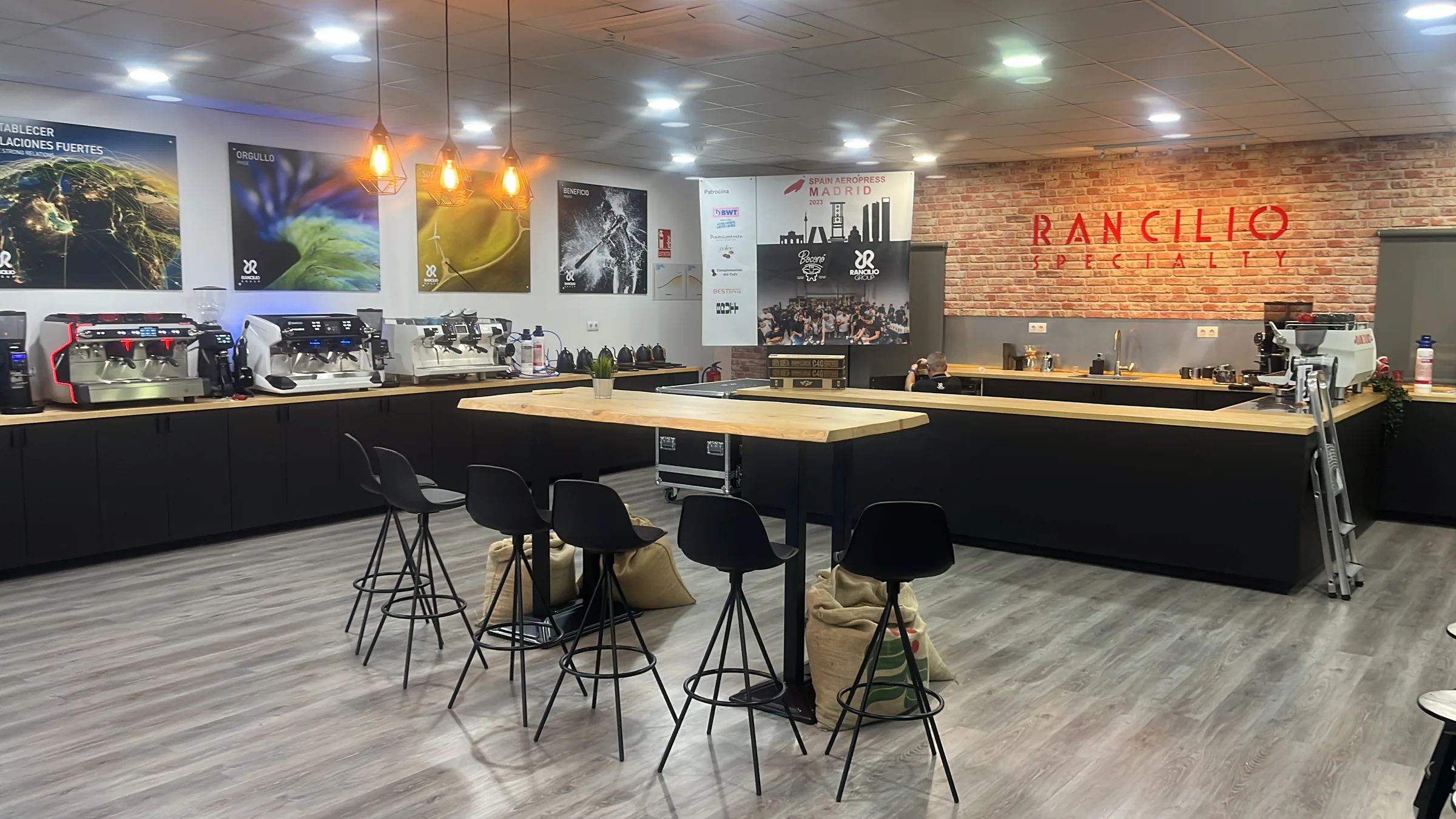 Showroom de Rancilio en Pinto con una mesa alta de madera, taburetes, máquinas de espresso y el rótulo «Rancilio Specialty».