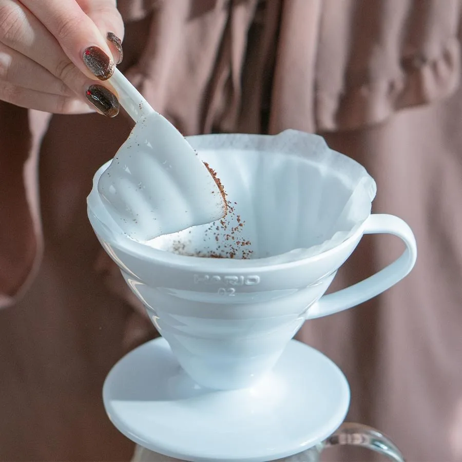 Una mano añade café molido con cuchara al Hario V60 02 blanco