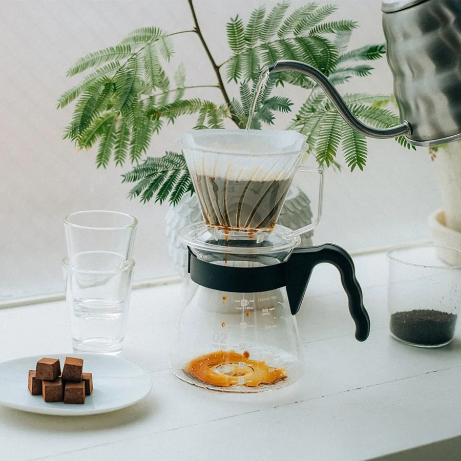 Hario V60 Coffee Server 700 ml bajo un gotero en una mesa con plantas