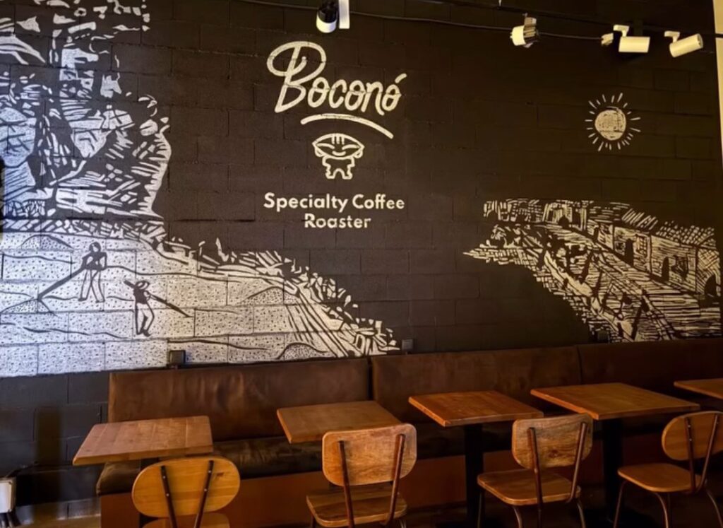 Mural blanco sobre la pared negra de la cafetería de Boconó en X-Madrid, con el logotipo de Boconó, escenas de trabajo en el campo y mesas de madera delante