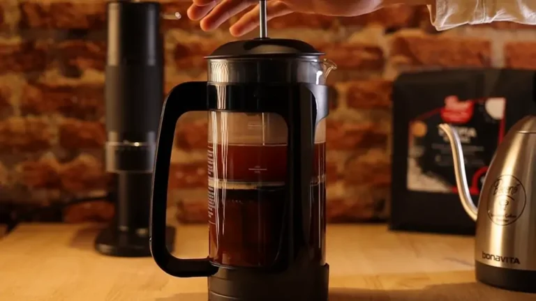 Prensa francesa con el café en infusión sobre una mesa de madera, con un molinillo y una bolsa de café de Boconó detrás
