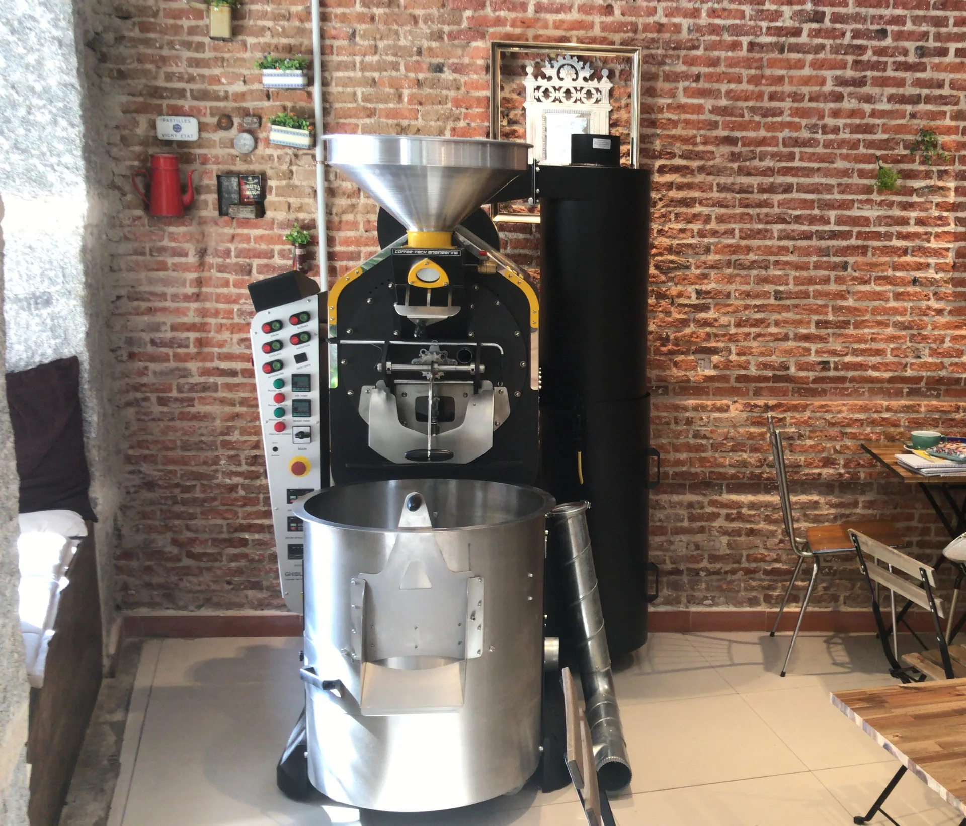 Tostadora de café Coffee-Tech con su tolva y la bandeja de enfriamiento, contra la pared de ladrillo y junto a las mesas de la cafetería de Boconó en Lavapiés, en 2018
