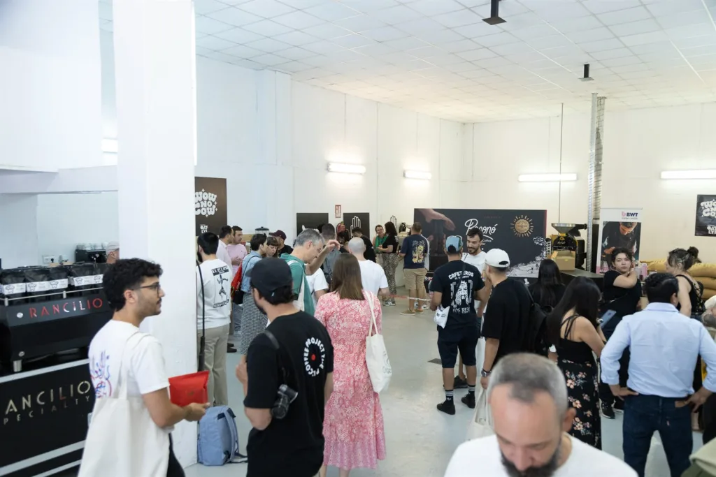 Nave del tostadero llena de gente, con sacos de café verde apilados y las mesas de competición al fondo