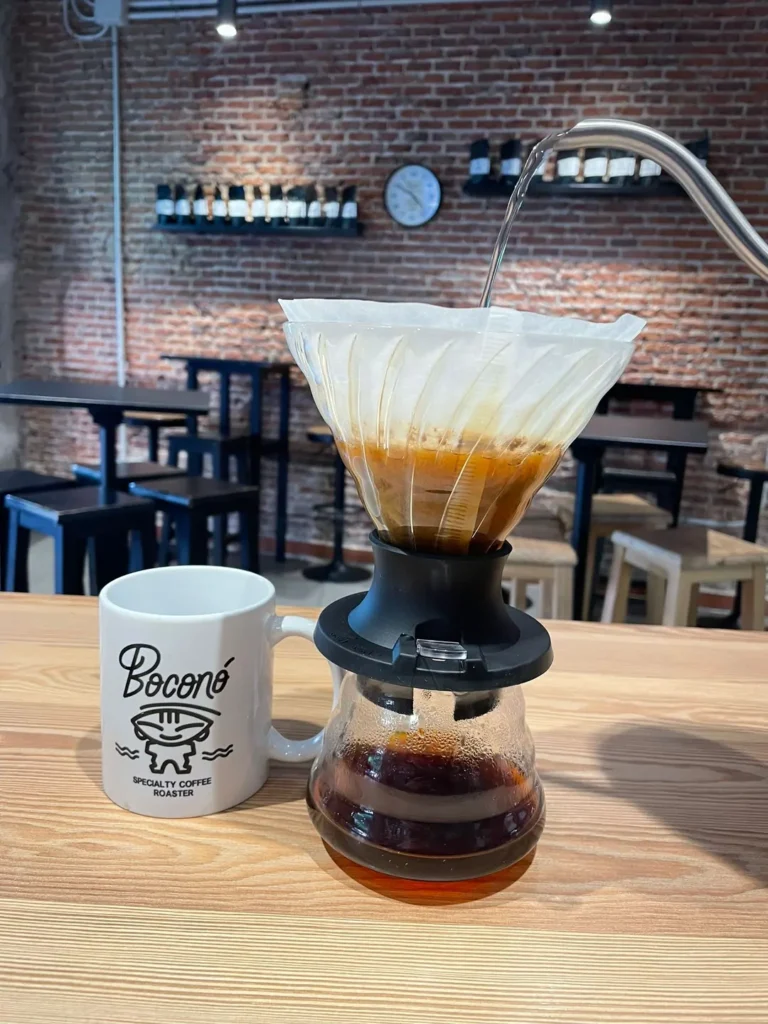 Café filtrándose en un V60 negro sobre una jarra de cristal, junto a una taza blanca con el logotipo de Boconó, en una barra de madera con una pared de ladrillo y estantes con bolsas de café detrás