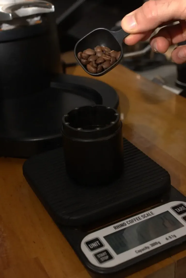 Una mano sostiene la cuchara con la dosis de café en grano junto a la báscula y el molinillo