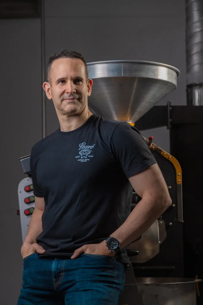 Carlos Núñez, tostador de Boconó y campeón Madrid Specialty Coffee Roaster 2023, junto al tostador de tambor del tostadero de Madrid, con las manos en los bolsillos